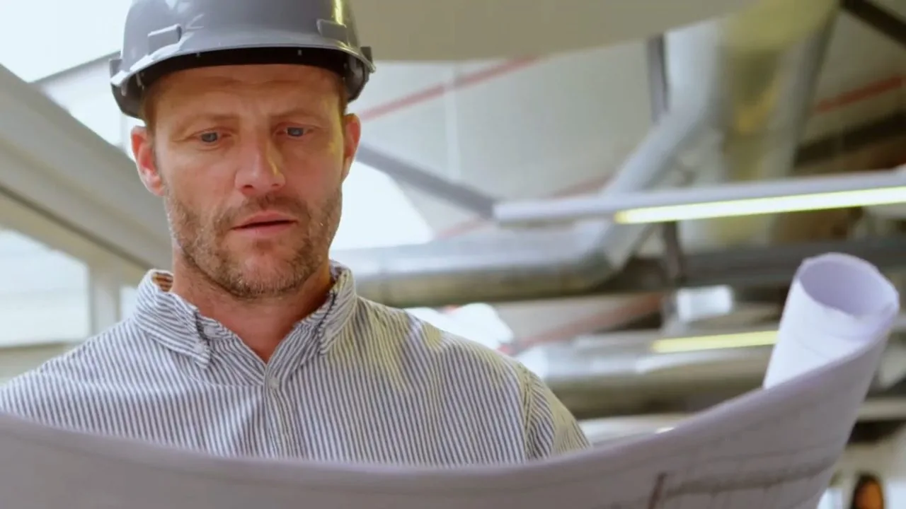 A man in a hard hat holding up printed plan sheets and looking at them.